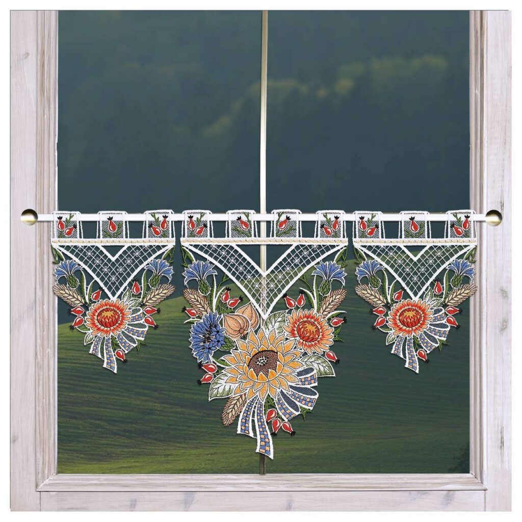 Ein Fenster mit einem Volant aus transparenter Spitze, geschmückt mit bunten Blumenstickereien in Gelb-, Blau-, Rot- und Grüntönen. Das komplizierte Muster zeigt große Blumen und Blätter mit einem geometrischen oberen Rand. Der Fensterrahmen ist aus Holz und draußen ist ein malerischer grüner Hügel zu sehen.