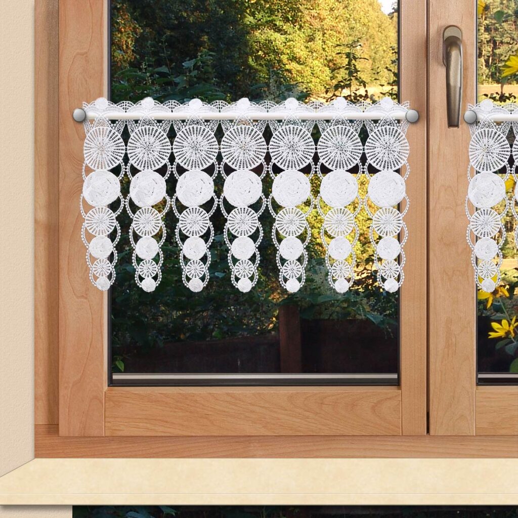 Ein Holzfenster mit Blick auf grüne Bäume und Sträucher im Hintergrund. Das Fenster ist teilweise mit einem weißen Spitzenvorhang bedeckt, der mit komplizierten kreisförmigen Mustern und herabhängenden Spitzenfransen verziert ist. Die Spitze lässt Licht durch und sorgt gleichzeitig für ein dekoratives Detail.